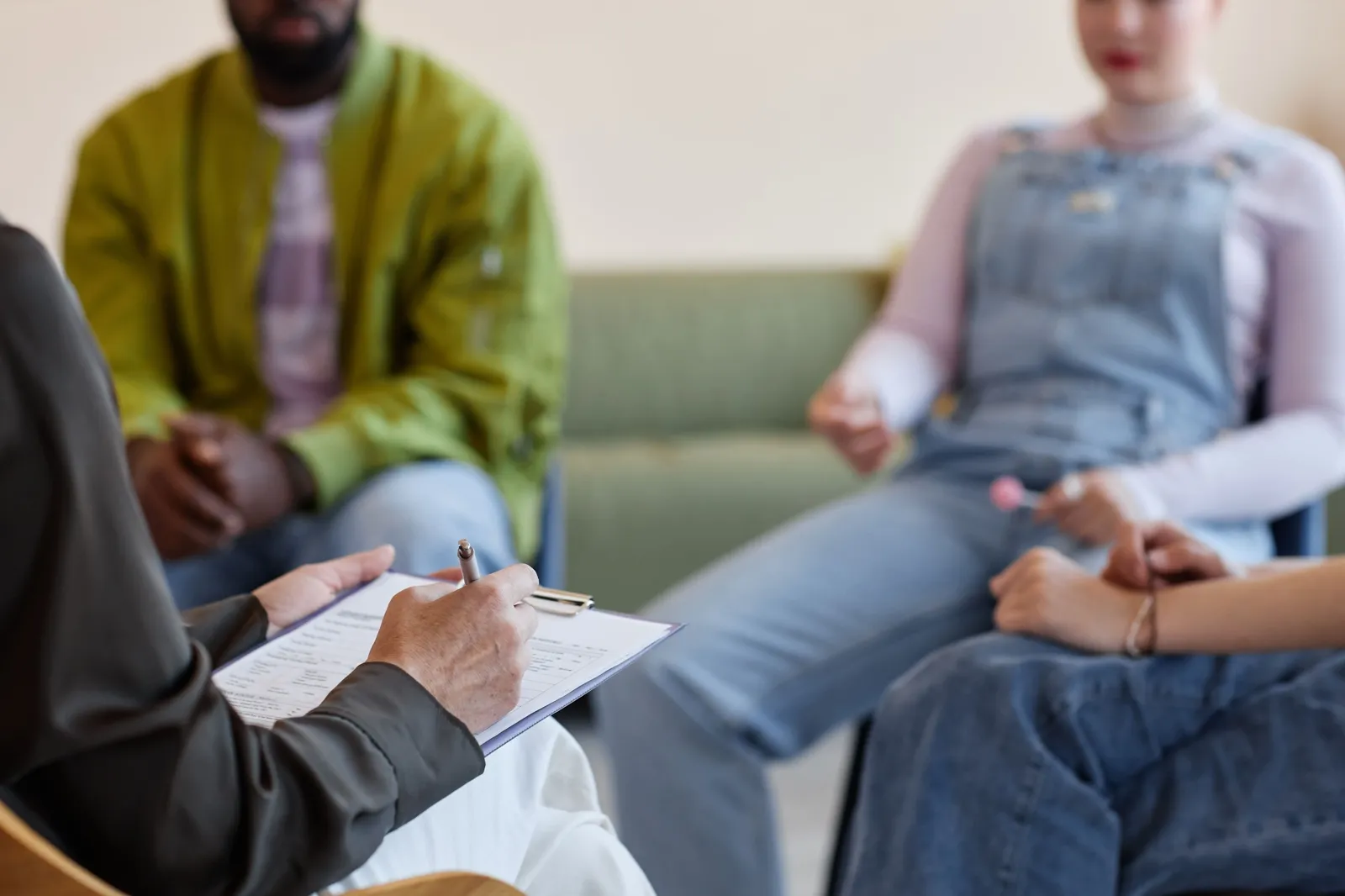 Close-up of a hand writing on a clipboard during a conversation, with two people seated on a sofa in the background