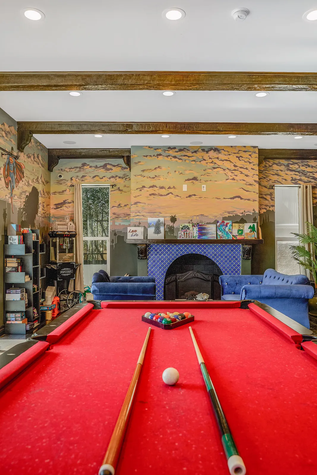 Red pool table beneath a beamed ceiling, with a painted mural and a tiled fireplace