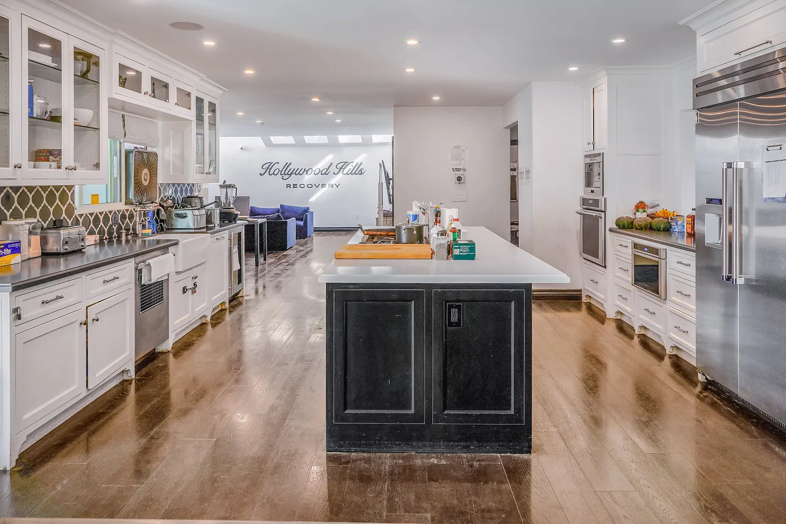 White kitchen with a long island, glass-fronted cabinets and the Hollywood Hills Recovery name on the wall
