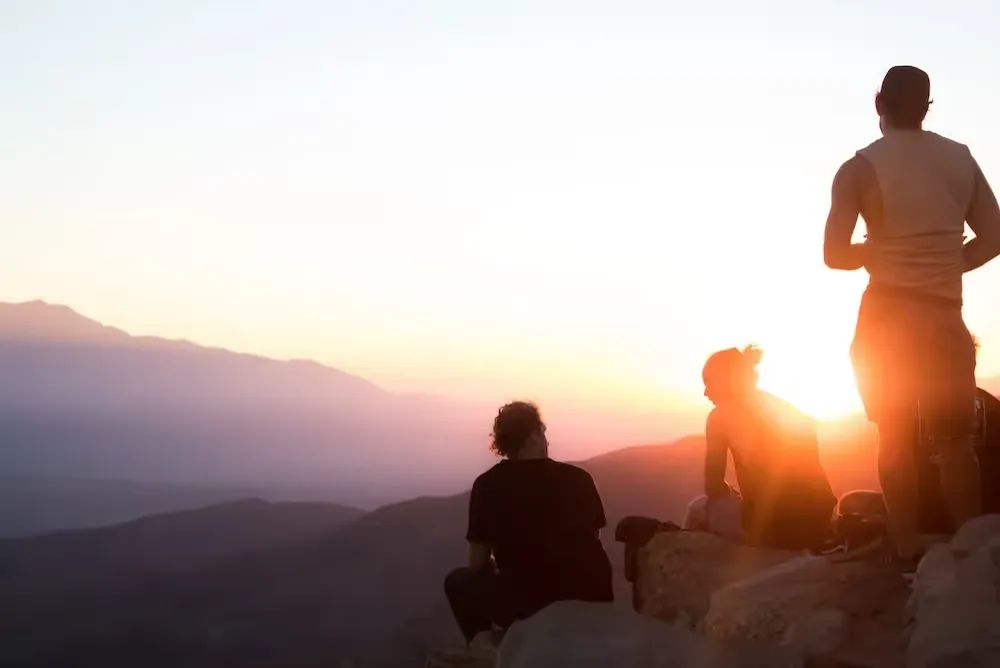 Three people on a rocky overlook watching the sun set over mountain ridges