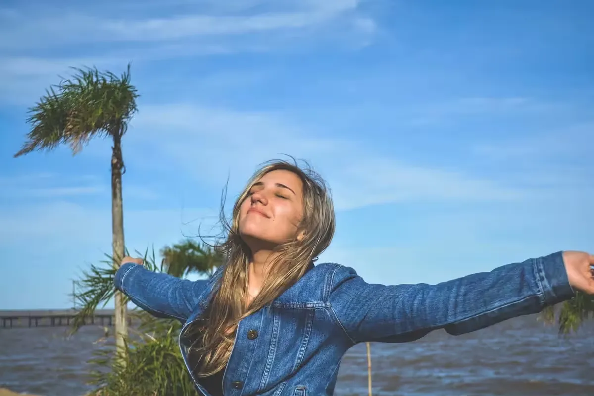 Woman in a denim jacket with arms outstretched beside the water on a sunny day, palm trees behind her